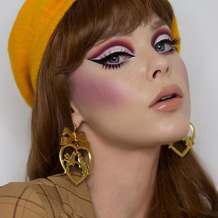 chica de cabello castaño claro usando maquillaje en colores morados, delineado tipo cat-eye, aretes dorados y boina amarilla