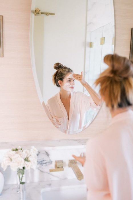 Chica con bata de baño sentada frente al espejo aplicando una mascarilla