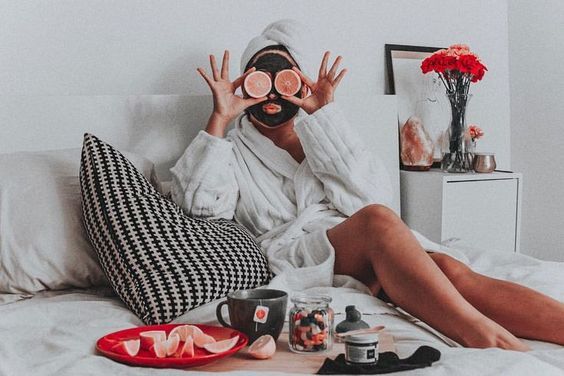 Chica sentada en su cama llevando bata de baño con mascarilla de arcilla negra y rodajas de toronja en el rostro