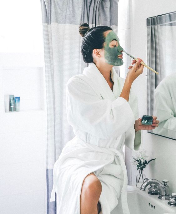 Chica con bata frente al espejo aplicando una mascarilla con brocha