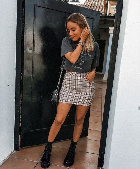 Chica rubia mirando hacia abajo tocándose la oreja y usando blusa gris, falda de cuadros y botas