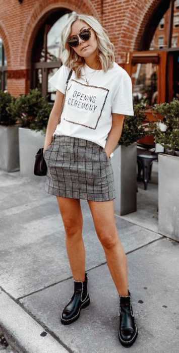 Chica rubia de cabello suelto pone las manos dentro de los bolsillos de su falda color gris con cuadros y usa una blusa blanca y botines negros