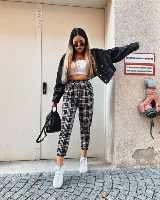 Chica estirando el brazo mientras sostiene una mochila y viste chamarra negra, blusa blanca y pantalón a cuadros blanco y negro