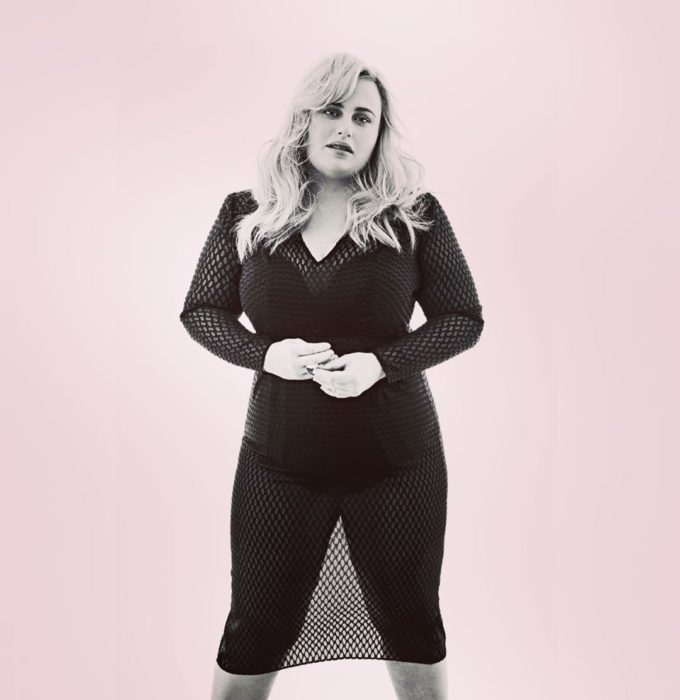 Rebel Wilson posando con ropa deportiva negra y un blusón estilo maya sobre un fondo rojo pastel