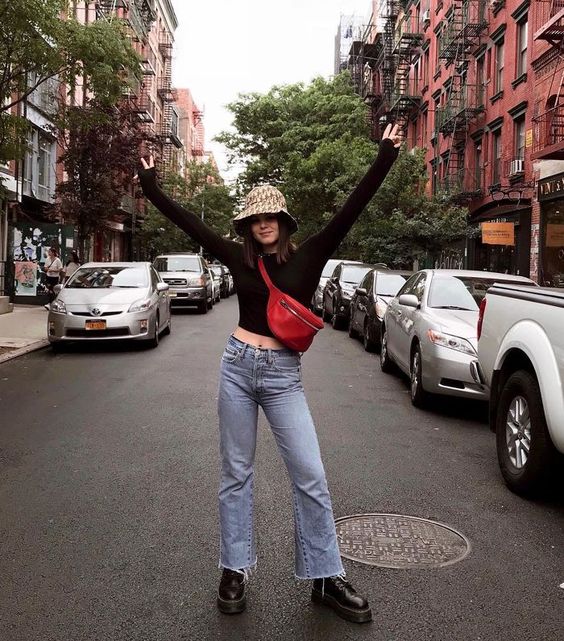 Chica parada a mitad de avenida llevando jeans de mezclilla, camisa negra y gorro estilo pescador