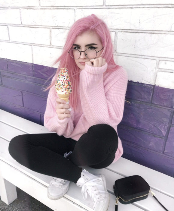 Cabello color rosa cherry blossom; chica sentada comiendo un helado con chispas de colores, suéter oversize tejido, lentes redondos vintage, cabello lacio y largo teñido