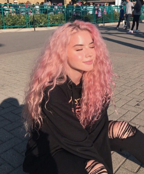 Cabello color rosa cherry blossom; mujer sentada bajo el sol, con los ojos cerrados, cabello largo y rizado