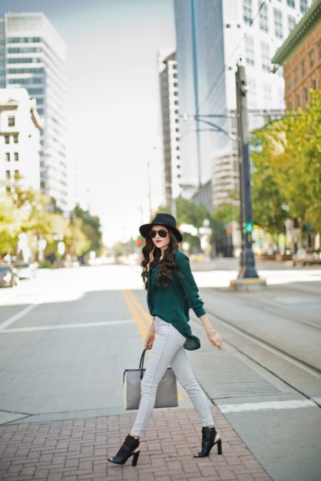 Chica elegante atraviesa la calle con pantalones de mezclilla claros, blusa verde esmeralda, sombrero negro y botines con abertura en la parte de adelante