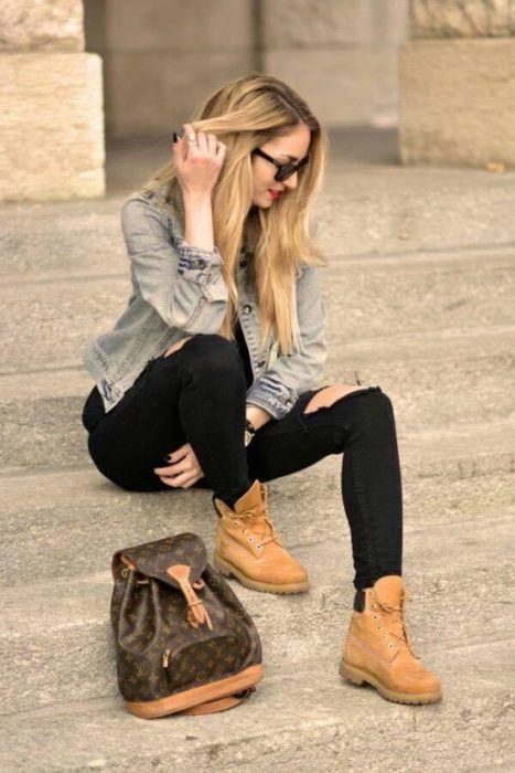 Mujer rubia sentada en escalones se toca el cabello con la mano derecha y viste pantalón negro, chamarra de mezclilla y botas tipo timberland color camel