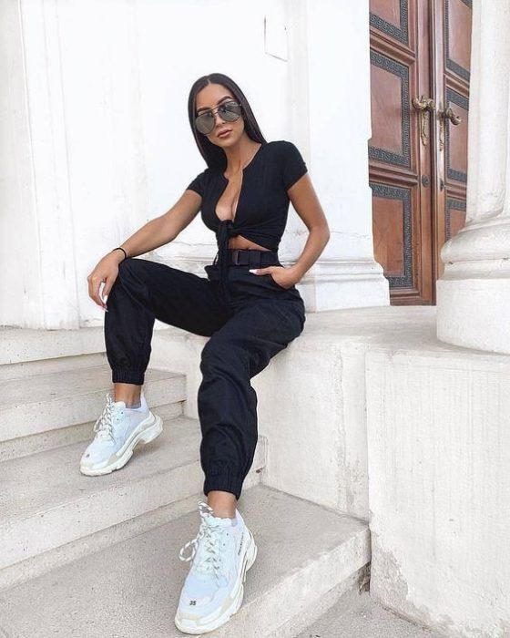 Chica con gafas de sol sentada en un escalón, vistiendo blusa negra y pantalón negro con tenis blancos