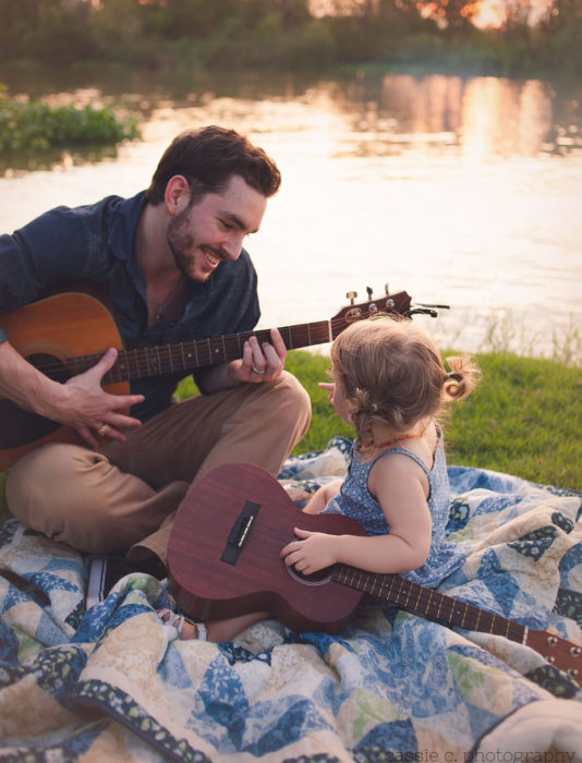 Frases del día del padre; papá tocando la guitarra con su hija en un día de campo al lado de un lago