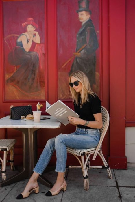 Mujer rubia sentada leyendo la carta de un restaurante, viste blusa negra, jeans, lentes de sol y zapatos cuadrados