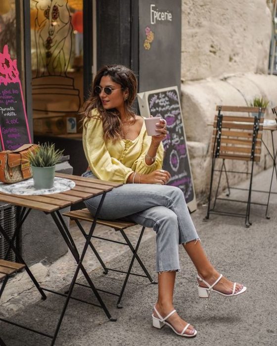 Mujer sentada, cruza la pierna, viste blusa amarilla, pantalón gris y tacones blancos cuadrados