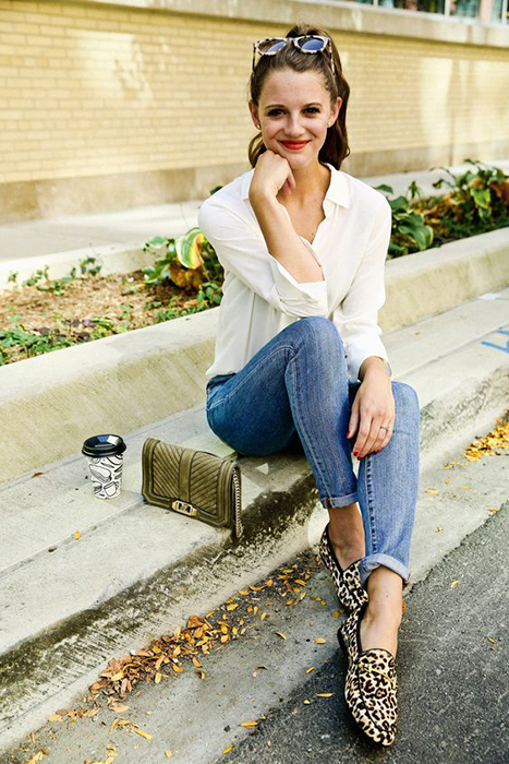 Mujer con blusa blanca, jeans y zapatos animal print sentada en la banqueta con su bolso a un lado