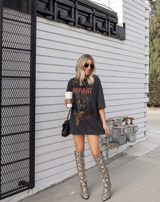Mujer rubia con melena corta y suelta posa con blusa amplia y larga negra con botas largas de animal print
