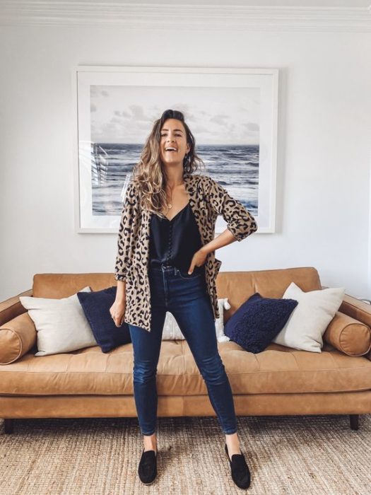 Mujer posa con mano en la cintura en la sala de su casa con saco de leopardo, blusa negra y jeans