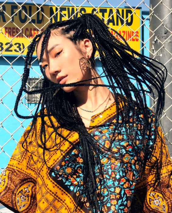 Looks con trenzas africanas; mujer asiática de ojos rasgados con cabello largo y negro, con peinado de coletas y aretes de rostro, ropa hippie