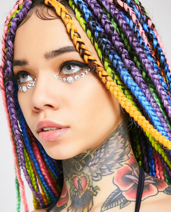 Looks con trenzas africanas; mujer de ojos grises, con cabello de colores arcoíris, anaranjado, verde, azul, morado y rosa, con tatuaje de búho, rosa y corazón en el cuello, con piedras en los ojos