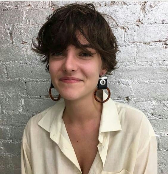 Chica sonríe y usa camisa blanca con cabello ondulado y muy corto y aretes largos