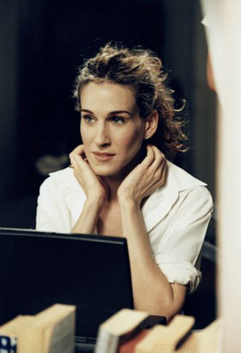 Carrie Bradshaw frente a su computadora usando una blusa blanca y cabello recogido en una coleta alta