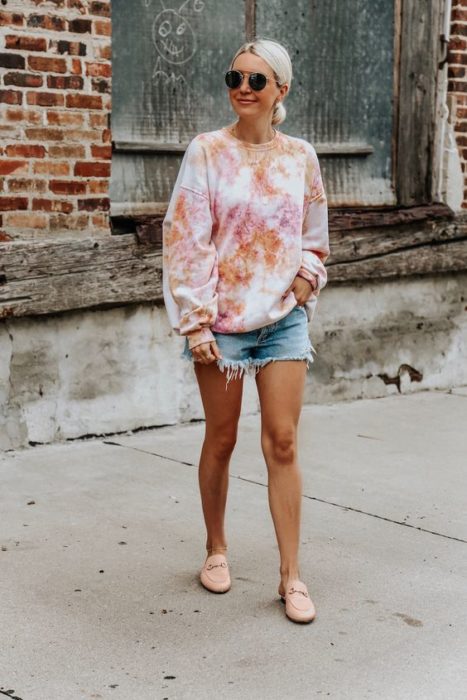 Chica rubia con cabello recogido posa con short de mezclilla y sudadera tie dye
