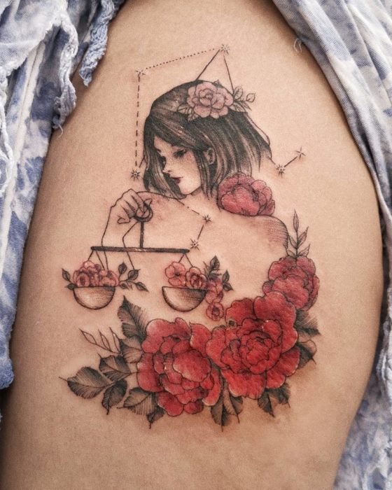 Tatuaje de una chica sosteniendo una balanza y flores a un lado, en tinta negra y color en la pierna