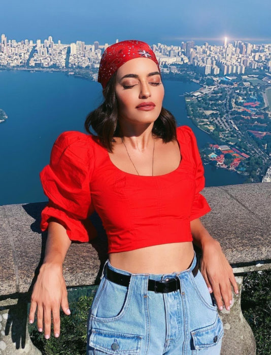 Chica disfrutando del sol con la ciudad de fondo, pañuelo en la cabeza y top rojo de mangas gigot