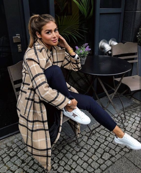 Chica sentada en silla usando un abrigo con estampado cuadrado, jeans azules, tenis blancos y coleta de caballo