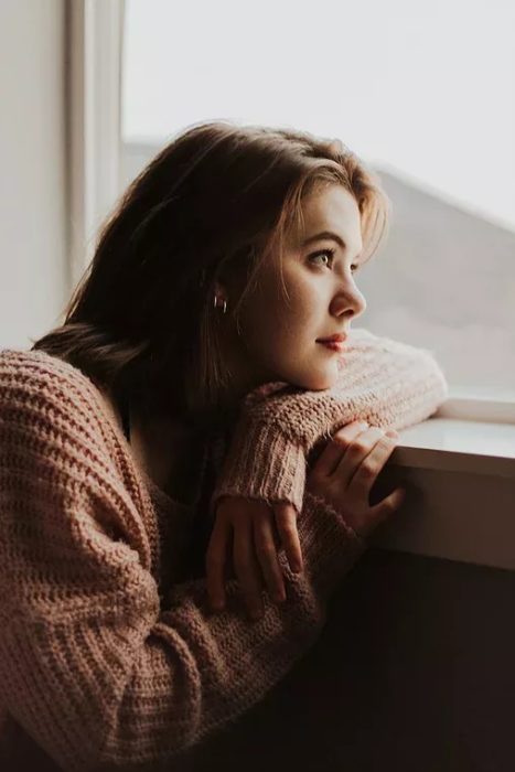 Mujer recargada sobre ventana mirando hacia afuera, vistiendo un suéter rosado