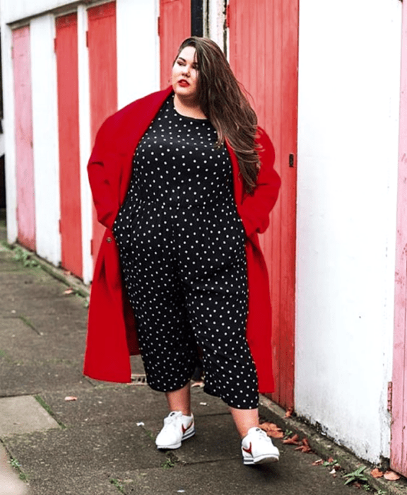 chica curvy de cabello castaño largo usando un jumpsuit negro de lunares blancos, tenis blancos deportivos y abrigo rojo largo
