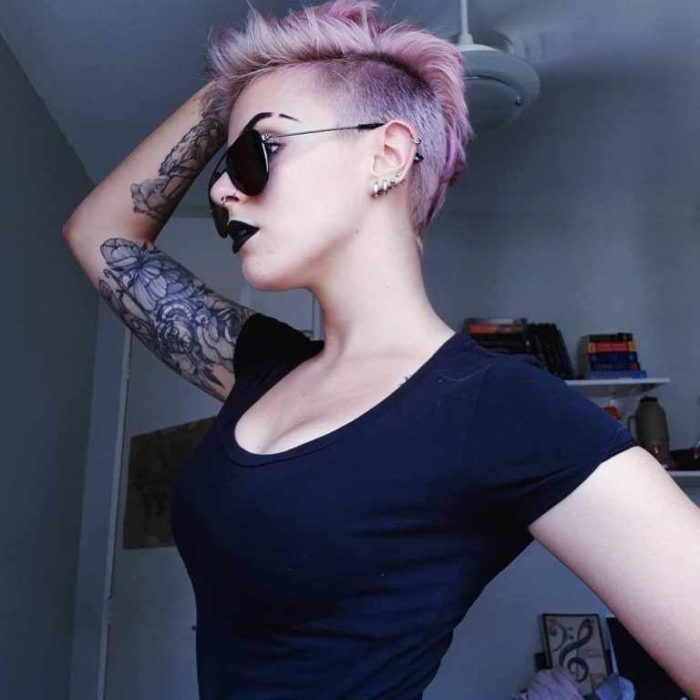 Modelo con corte de cabello rapado en tonos violeta, usando gafas de sol negras y blusa del mismo tono