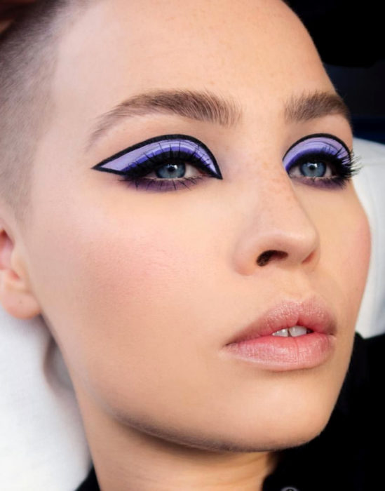 Maquillaje tendencia para el 2020; delineado gráfico o flotante con sombra morada; mujer rapada con pecas y teeth gap
