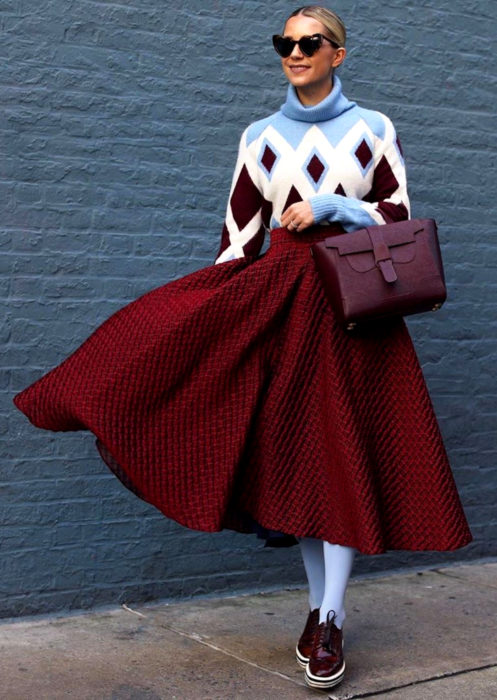 Outfits con medias de colores; mujer rubia con lentes de sol en forma de corazón, suéter oversized de rombos, falda midi rosa, medias azules y zapatos de charol color vino