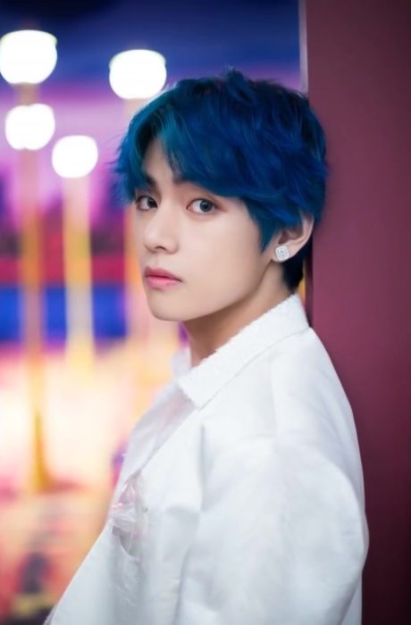 V con cabello azul rey recargado en una pared vino