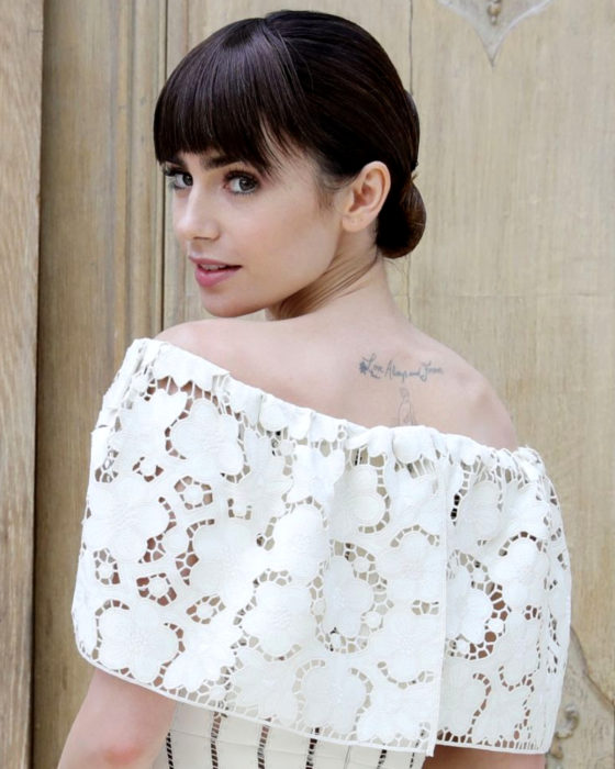 Peinados de Lily Collins para Año Nuevo; mujer de cabello castaño agarrado en un chongo bajo, con flequillo disparejo y tatuaje en la espalda