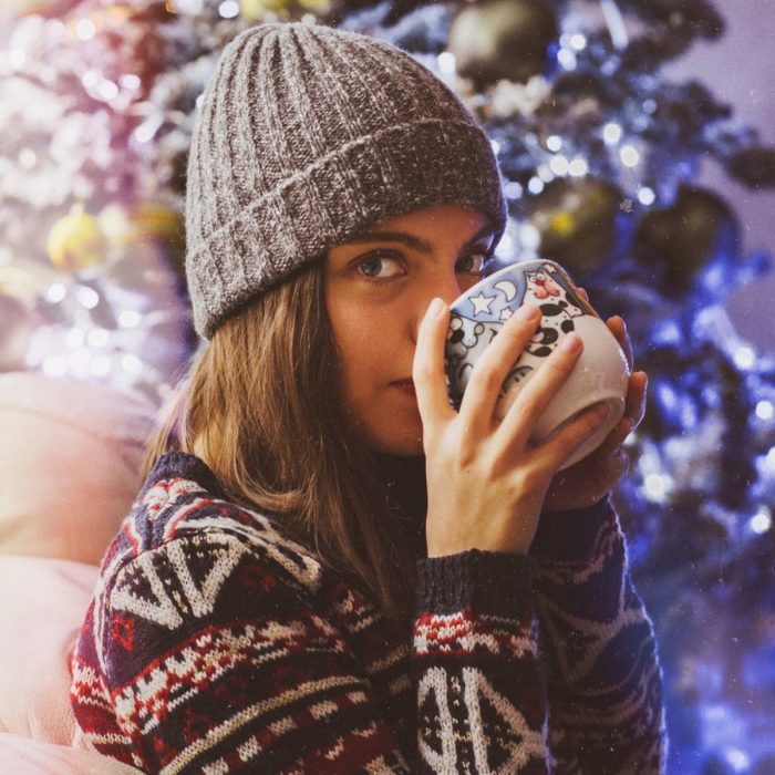 Chica con suéter navideño y gorro tejido, bebiendo café en una taza de gatos frente al árbol de Navidad con luces azules