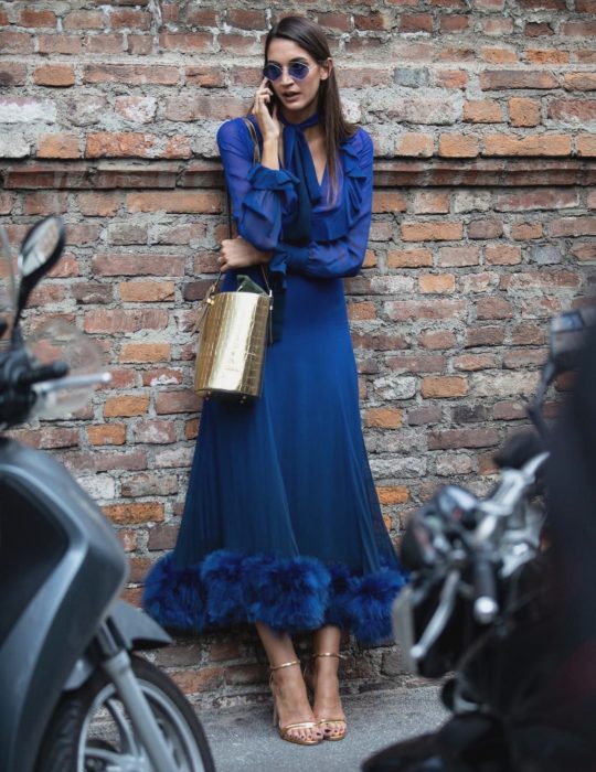 Pantone elige el classic blue como el color del 2020; mujer recargada en la pared, hablando por celular, con vestido azul clásico y bolsa dorada