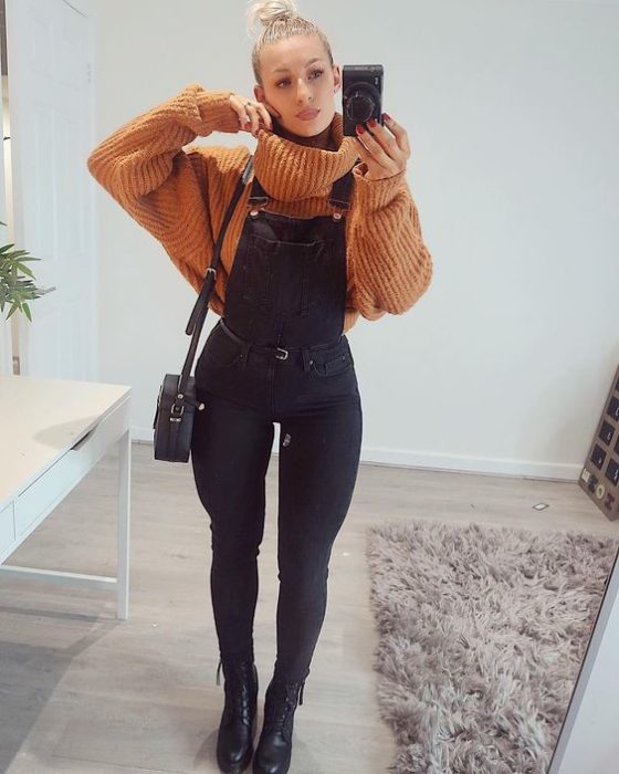 Chica tomándose una selfie mientras usa un overol negro con un suéter de color naranja