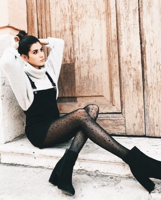Chica sentada afuera de una casa mientras posa con un overol negro, suéter blanco y botines