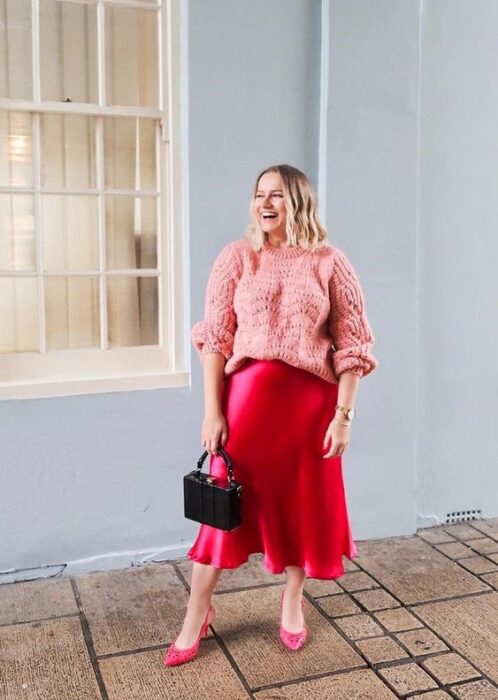 Chica plus size con cabello suelto ondulado vistiendo una falda de satín roja, suéter rosa y bolso negro