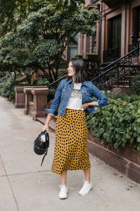 Chica de cabello corto con blusa blanca, chaqueta de mezclilla y maxi falda amarilla con puntos negros y tenis blancos