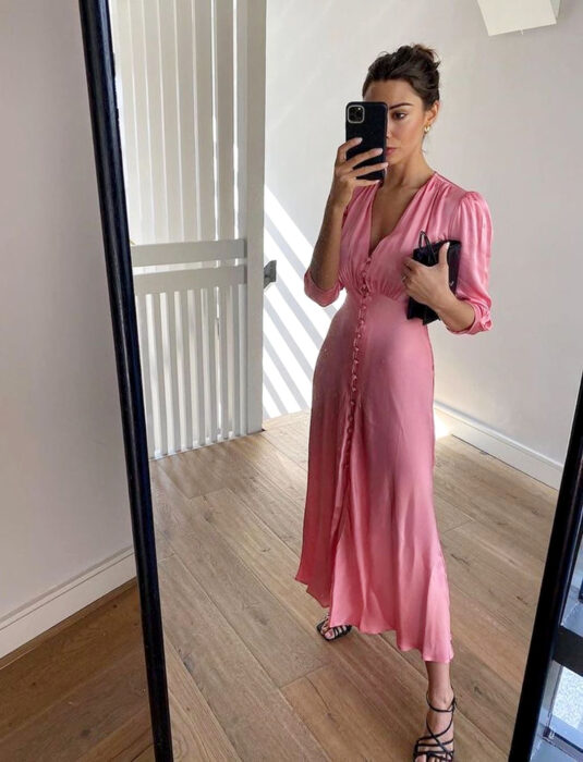 Mujer tomándose una selfie con su celular frente al espejo, vestido rosa largo y liso, de mangas a los codos, escote en v y botones al frente, peinado de chongo recogido y cabello castaño, bolsa de mano negra, sandalias de tacón negras