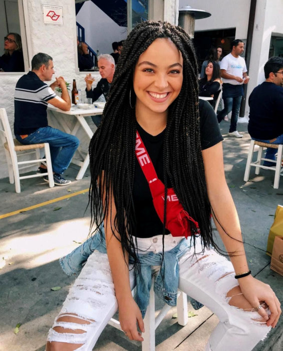 Looks con trenzas africanas; mujer sonriendo sentada en un café, cabello largo y negro, jeans desgastados