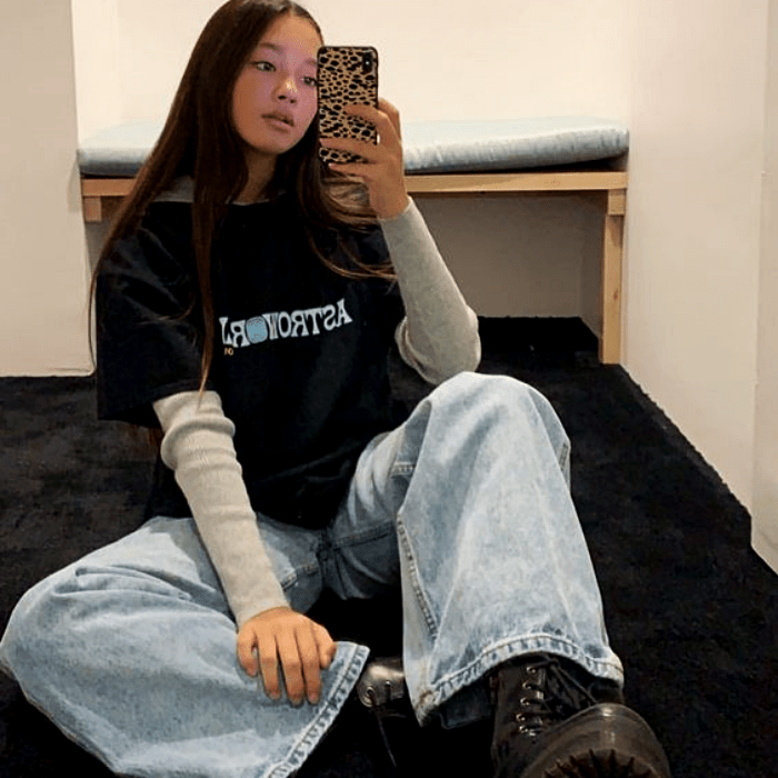 chica de cabello largo castaño usando una camiseta negra de astroworld con mangas grises, pantalón de mezclilla baggy y botas negras de plataforma