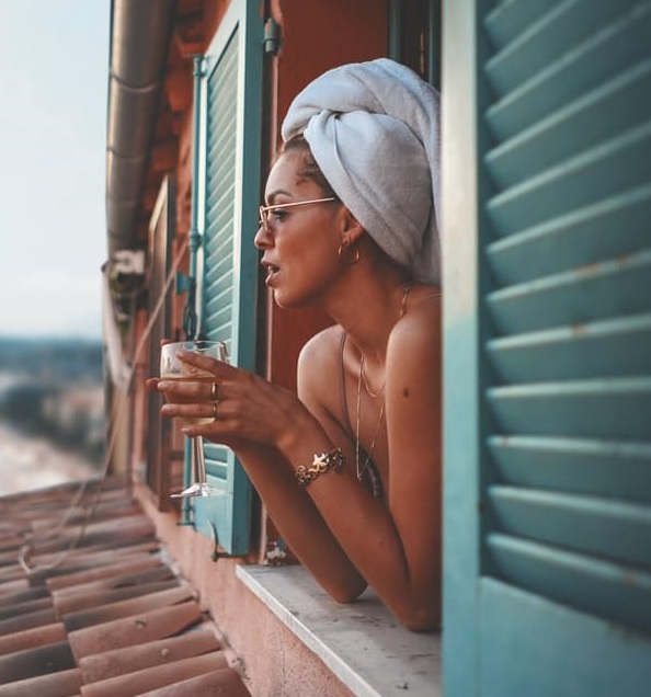 Chica con toalla blanca en la cabeza asomándose por la ventana con una copa de vino blanco