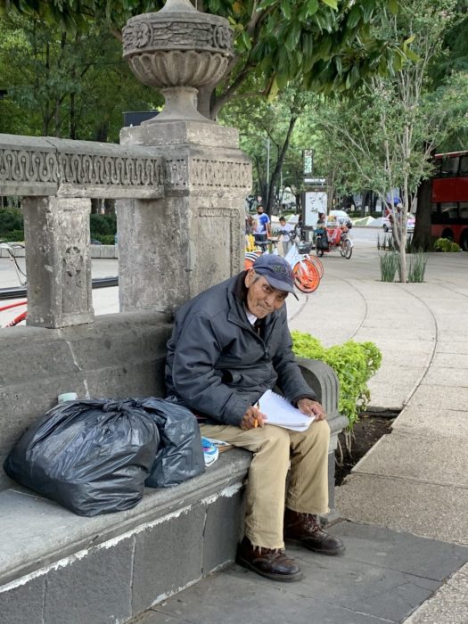 Don Baldemar, abuelo sentado en una banca de cemento dibujando en una libreta blanca, vende dibujos para sobrevivir