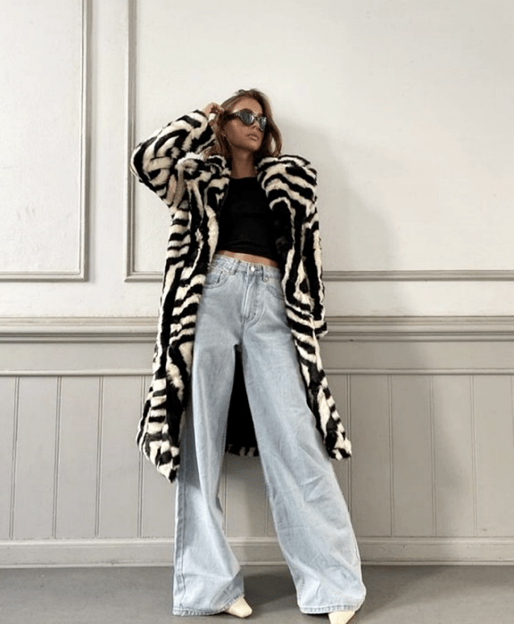 chica rubia usando lentes de sol, top negro, abrigo afelpado de cebra, jeans acampanados a la cintura y botines blancos de piel