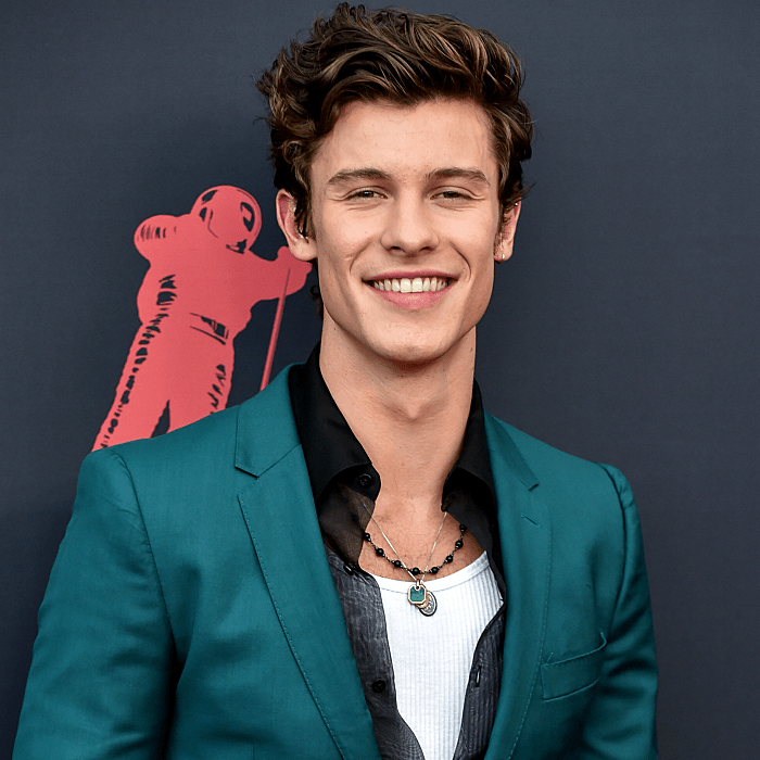Shawn Mendes usando una camiseta de tirantes blanca, camisa negra semitransparente y saco blazer color turquesa
