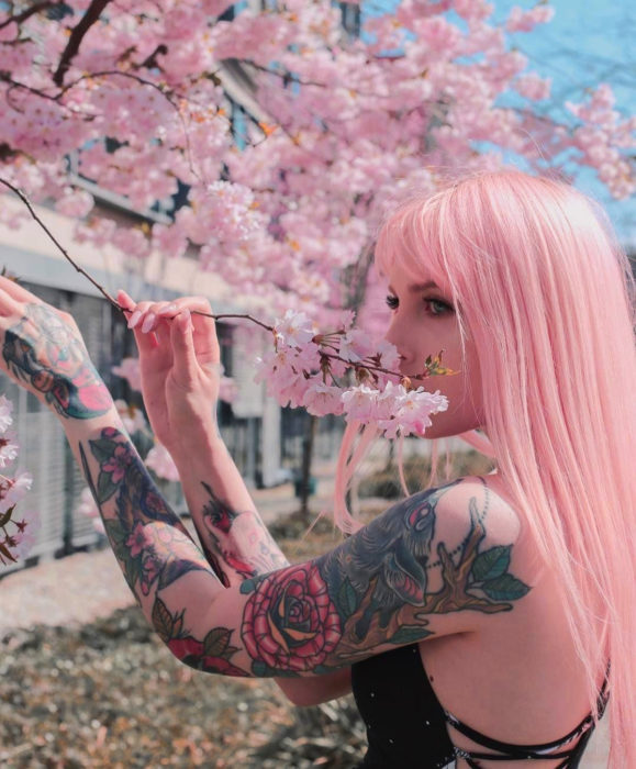Cabello color rosa cherry blossom; mujer con tatuajes en los brazos junto a un árbol de flores rosas, cabello lacio y largo