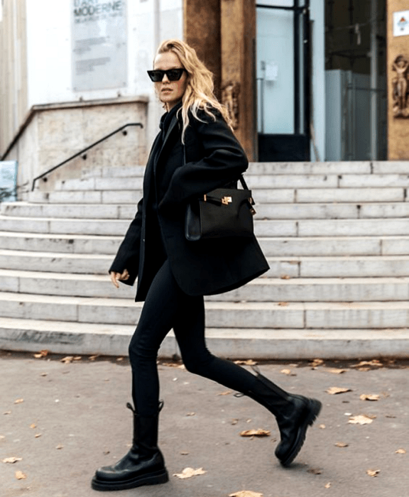 Chica rubia usando lentes de sol, abrigo negro, leggings negros, botines de plataforma negros de piel y bolso negro de piel